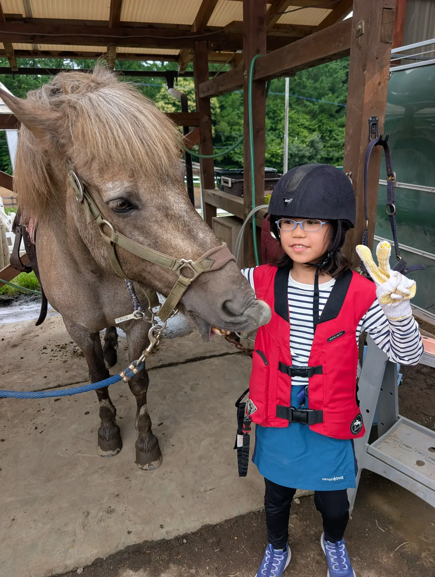 Child em um riding helmet e vermelhos segurança vest beside um saddled pony em um covered wooden shelter
