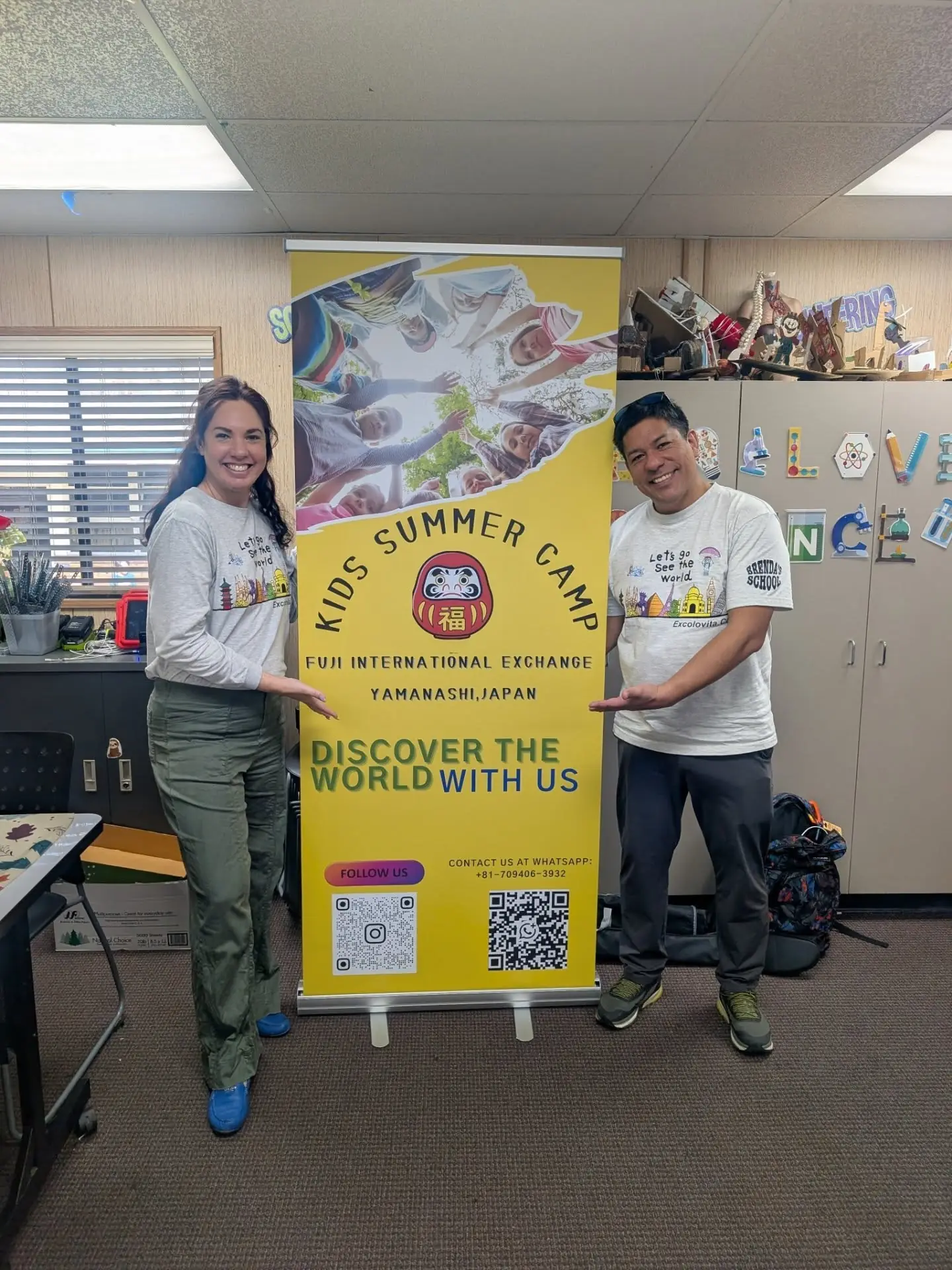 Two adults standing beside a yellow Fuji International Exchange banner indoors.